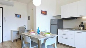A compact kitchen with white cabinets, a refrigerator, and a dining area with a table set for two. - Hostdomus - Castelli Apartment (Finale Ligure)