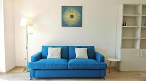 A blue sofa with two pillows, a floor lamp, a bookshelf, and a painting on the wall. - Hostdomus - Castelli Apartment (Finale Ligure)