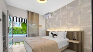 A modern hotel room with a bed, a bedside table, an air conditioning unit, and a view of a garden through a glass door.