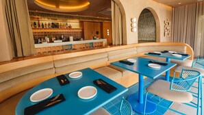 A restaurant with blue tables and chairs, a bar area with shelves of bottles, and a seating area with cushioned benches.