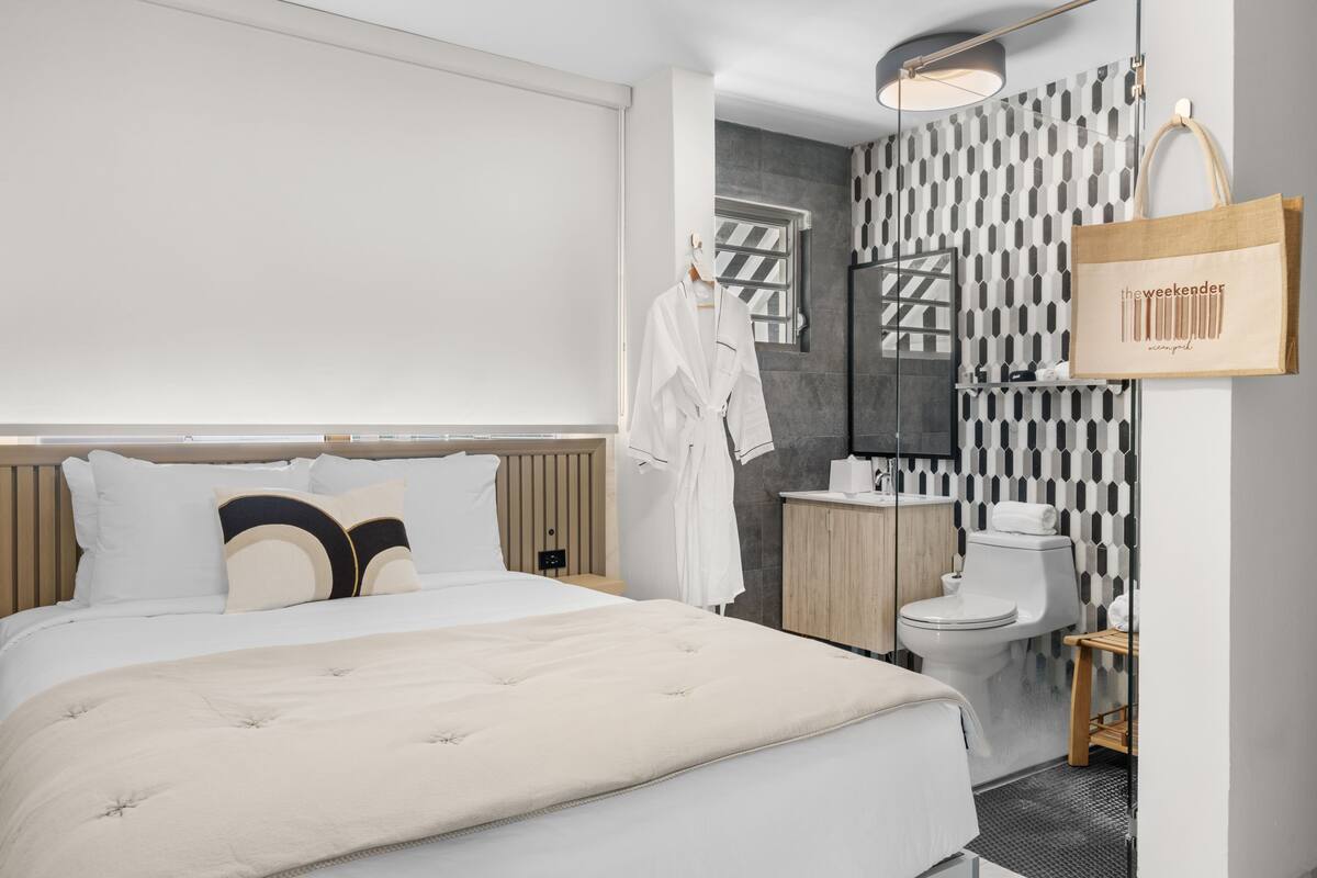 Habitación de hotel moderna con una cama grande, cabecera de madera, baño con pared estampada y un bolso colgado.