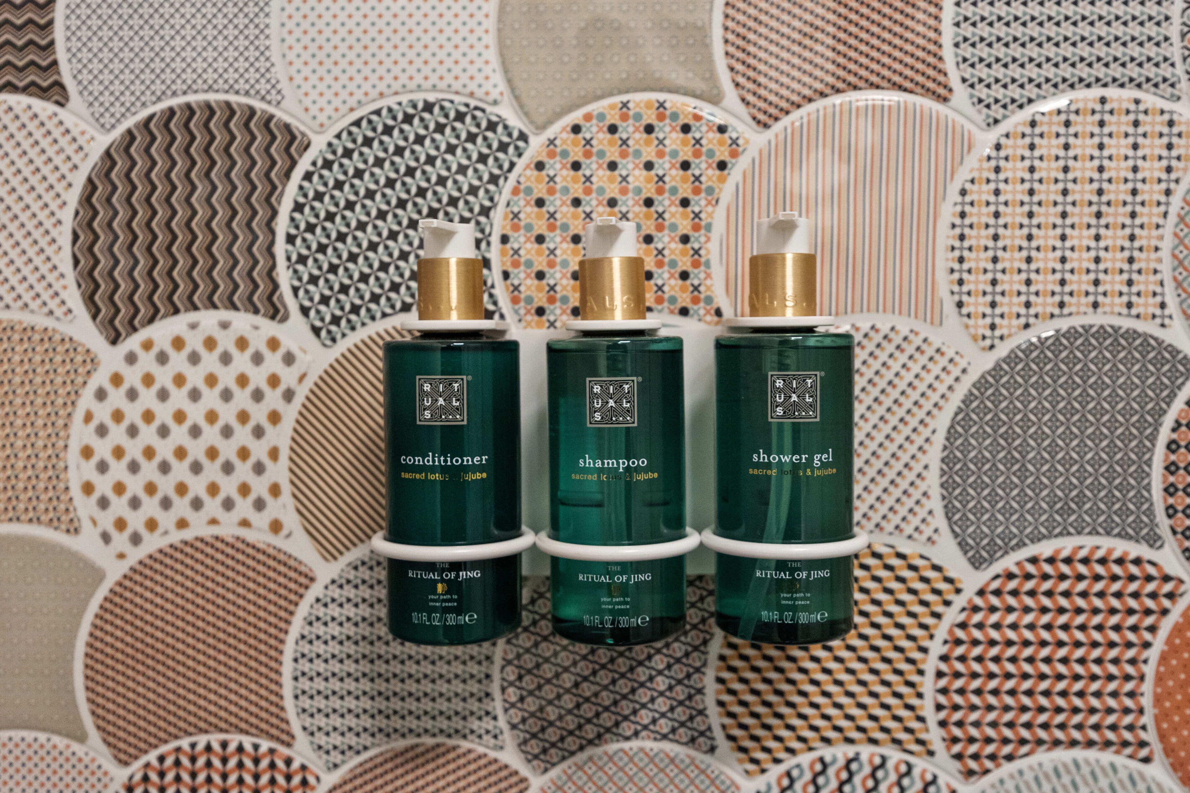 Tres botellas de productos Ritual de Jing: acondicionador, champú y gel de ducha, dispuestas sobre una superficie estampada.