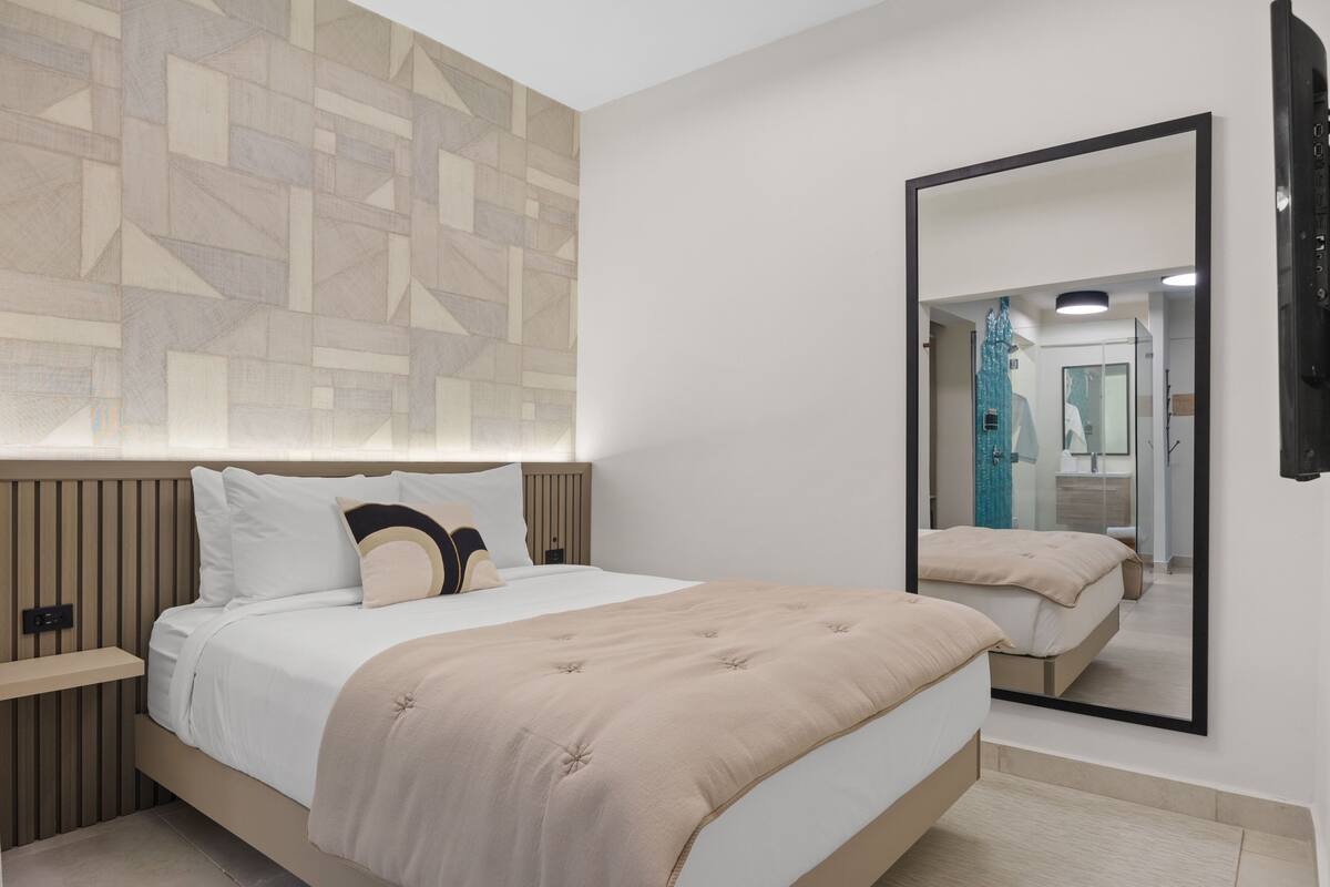 Habitación de hotel moderna con una cama grande, una mesita de noche, un televisor montado en la pared y un espejo que refleja la habitación.