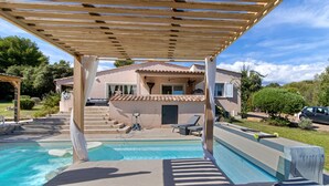Zona de piscina con una pérgola, sillones de descanso y una casa al fondo.