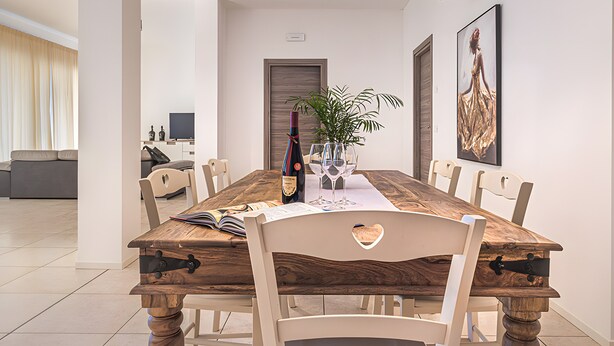 A dining area with a wooden table, white chairs, a potted plant, and a wine bottle.