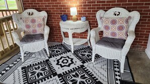 A cozy seating area with two white wicker chairs, a small table with a lamp, and a patterned rug. - Charming, convenient, & affordable house near Flint. Sleeps 12 comfortably! (Bainbridge)