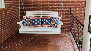 A white swing suspended by chains against a brick wall, a white column, and a tiled floor. - Charming, convenient, & affordable house near Flint. Sleeps 12 comfortably! (Bainbridge)