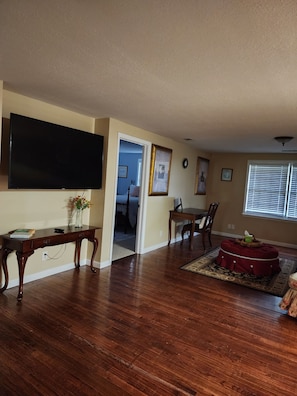A living room with hardwood flooring, a flat-screen TV mounted on the wall, a dining table with chairs, and a red ottoman.