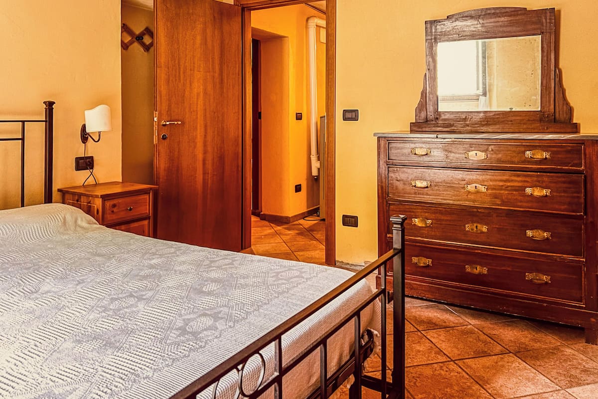 A bedroom with a bed, wooden dresser, nightstand, and a wall-mounted lamp.