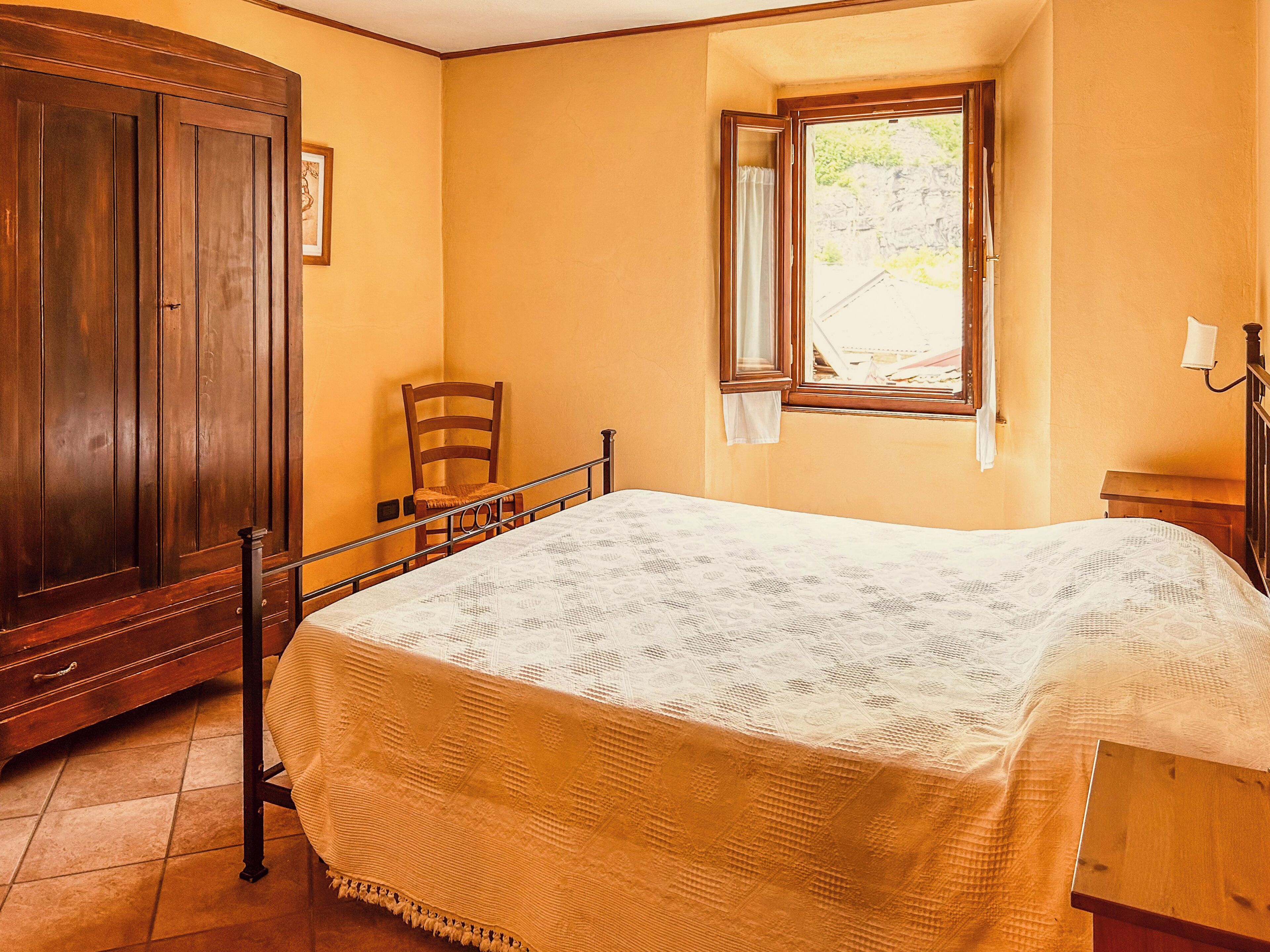 A bedroom with a bed, wooden wardrobe, chair, window, and lamp.
