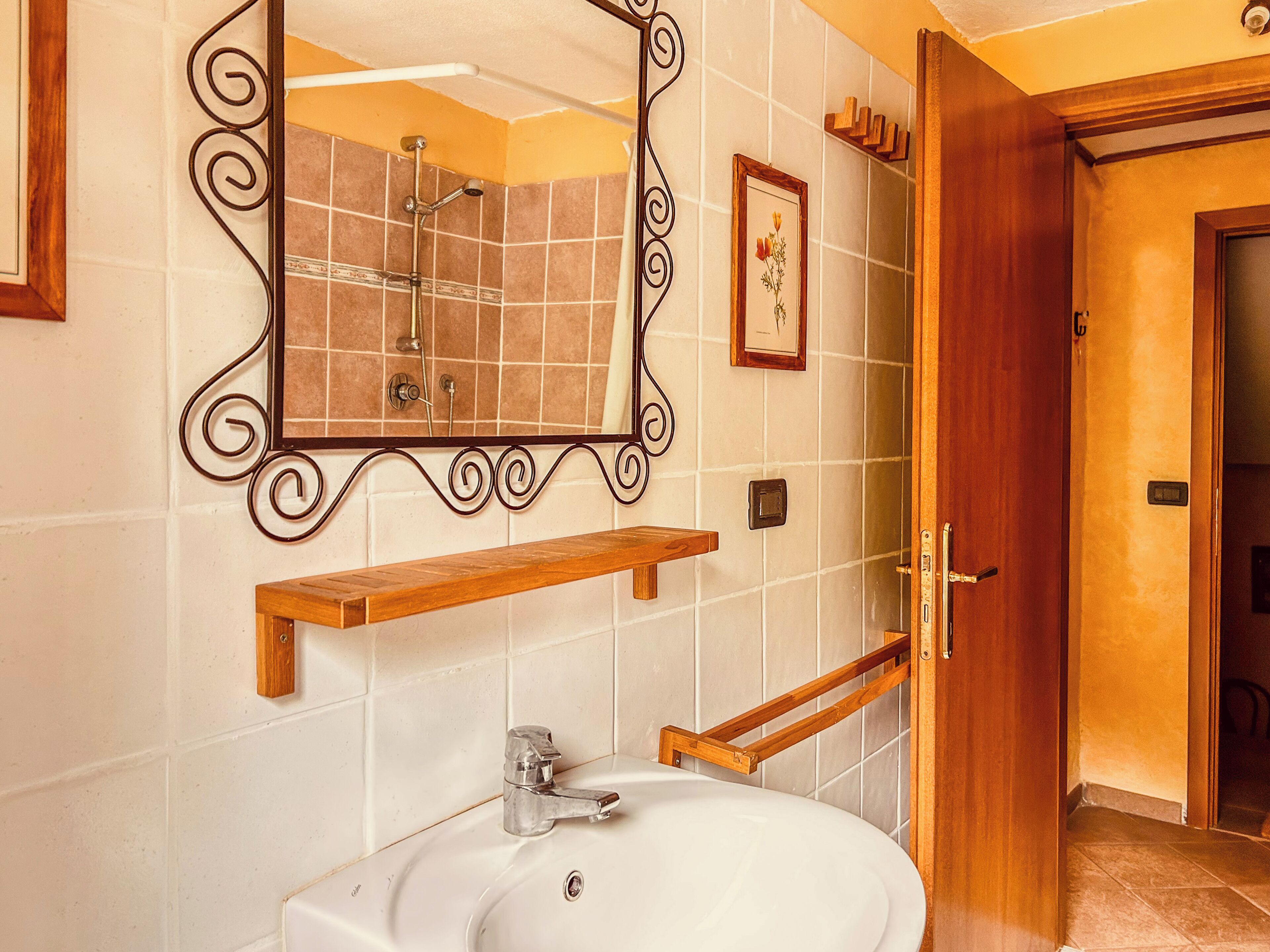 A bathroom with a white sink, a shower, a mirror with a decorative frame, and a wooden shelf.