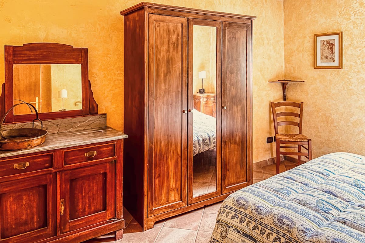 A bedroom with a wooden wardrobe, a dresser, a bed with patterned bedding, a chair, and a framed picture on the wall.