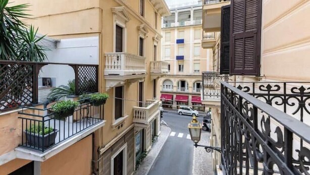 A narrow street with buildings on either side, balconies with plants, and a street lamp. - Hostdomus - Cherry Home (Alassio)