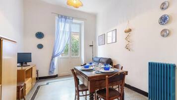 A dining area with a table set for two, a television on a stand, and a window with curtains.