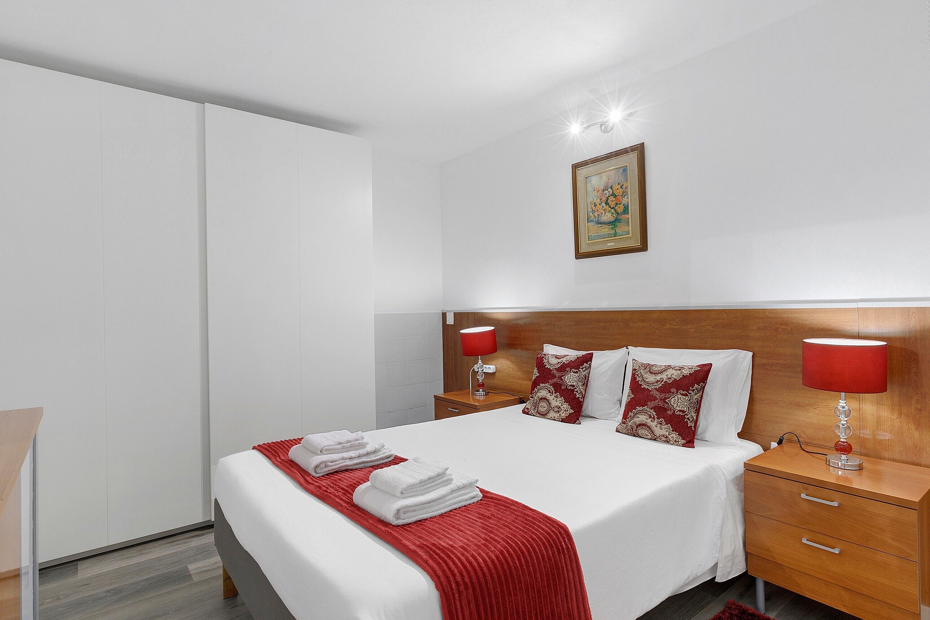 Cama bem arrumada com lençóis brancos e um cobertor vermelho, mesa de cabeceira de madeira com luminária vermelha e cabeceira de madeira.