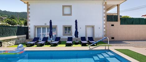 Piscina com espreguiçadeiras e guarda-sóis, um edifício branco com telhado em azulejos e um viveiro verde.
