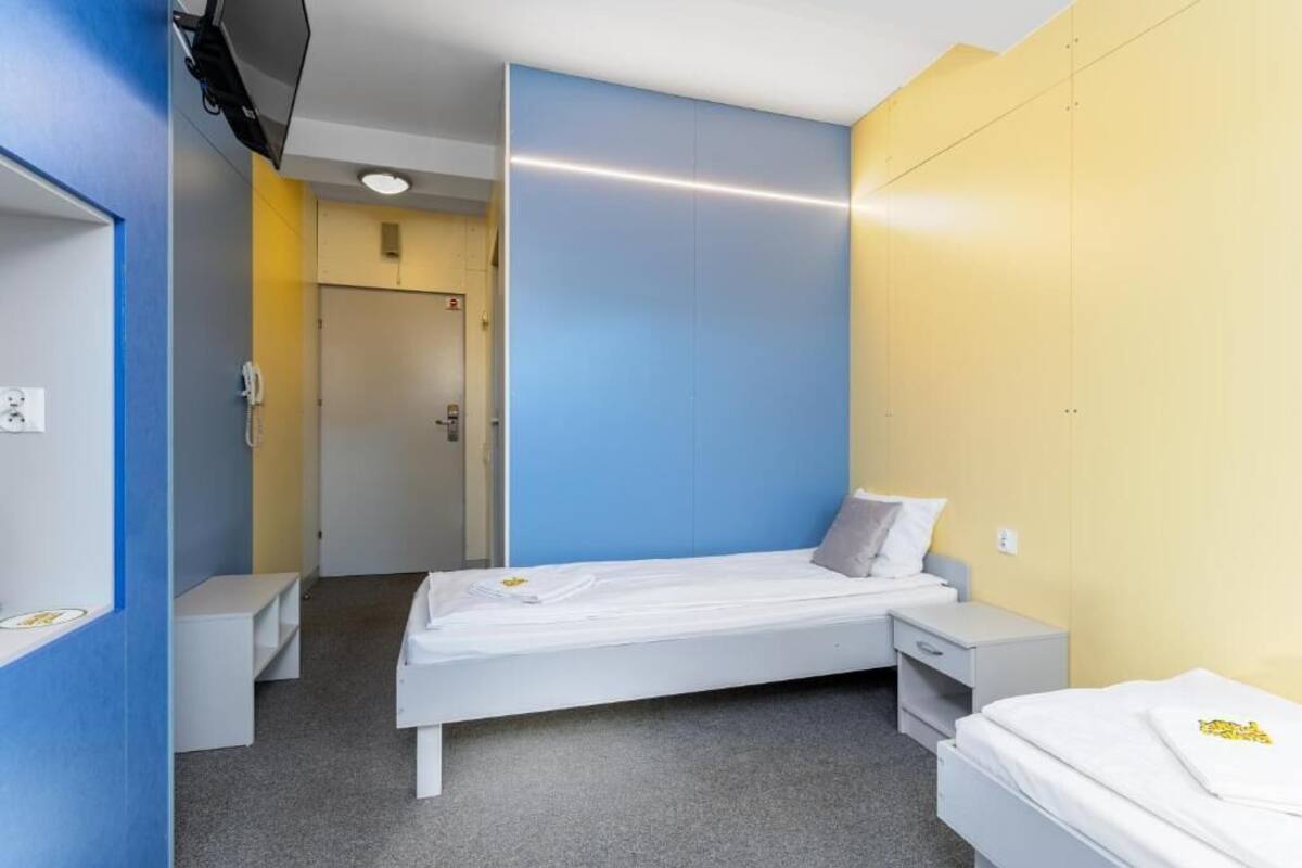 A single-bed room with a grey carpet, a white bed, a small bedside table, a wall-mounted TV, and a door leading to another room.