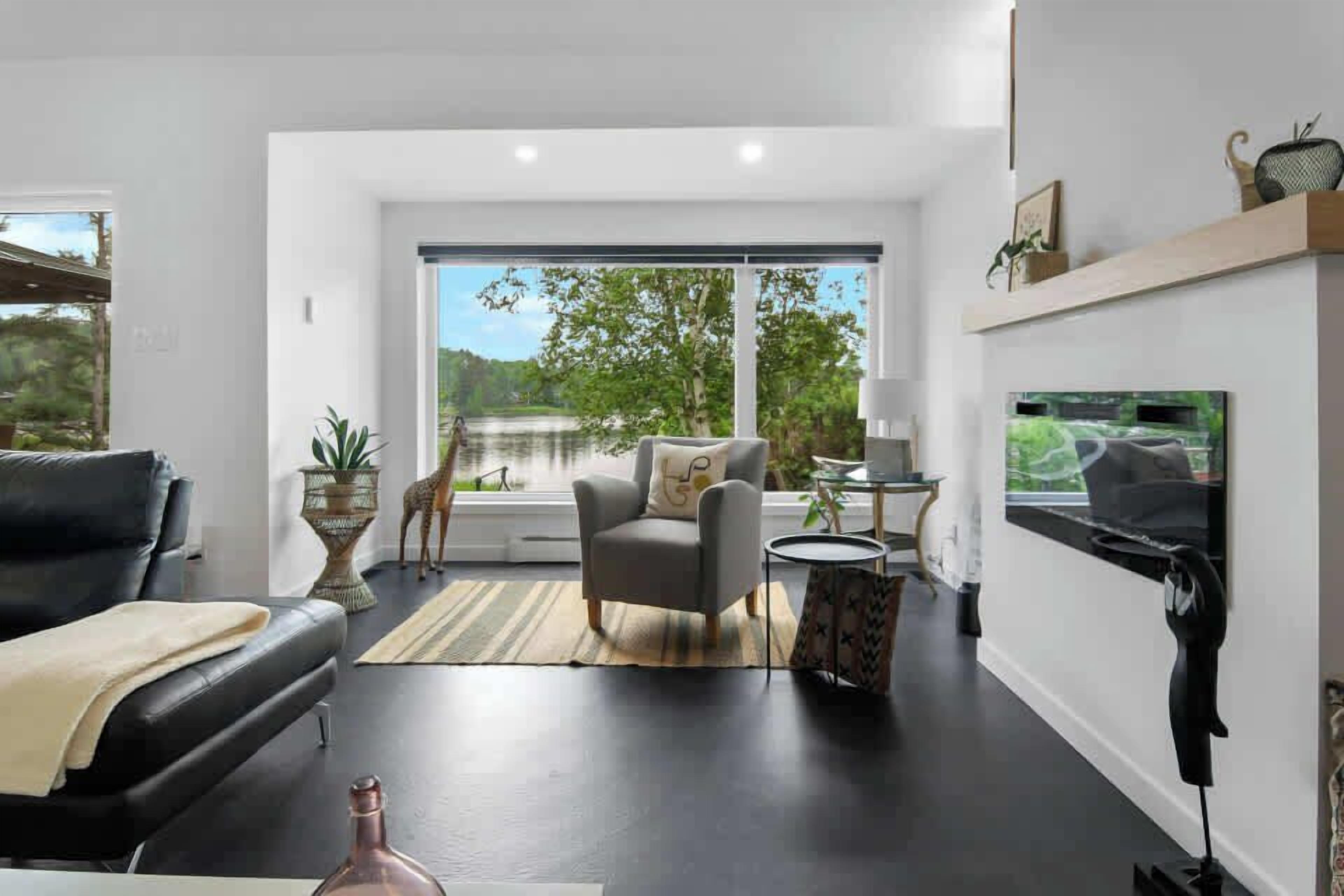 Un salon moderne avec un canapé en cuir noir, un fauteuil gris, une petite table ronde, un téléviseur fixé au mur et une grande fenêtre donnant sur un paysage verdoyant.