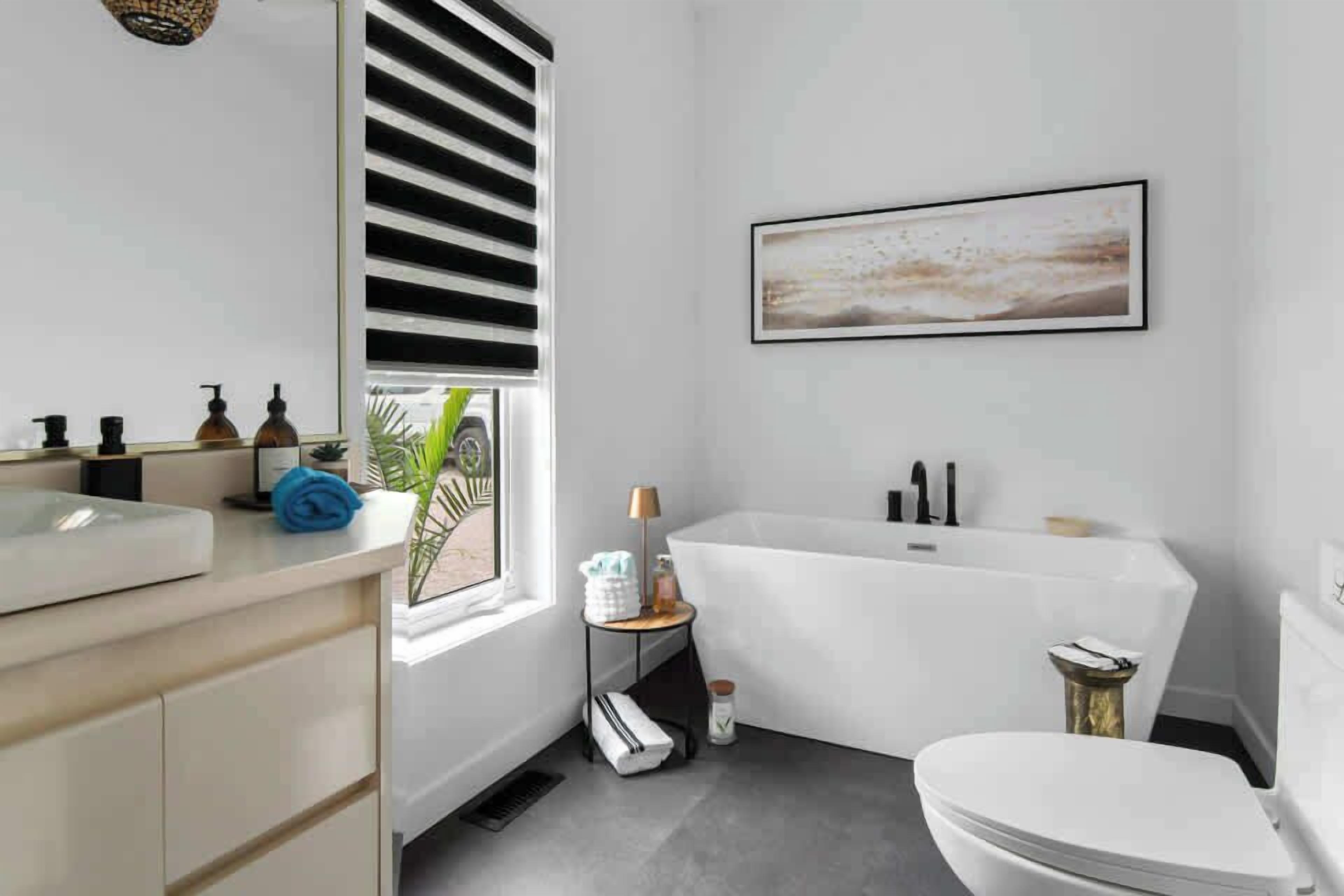 Une salle de bain moderne avec une baignoire, des toilettes et un lavabo. On y trouve une fenêtre avec des stores, un porte-serviettes et une petite table avec une lampe.