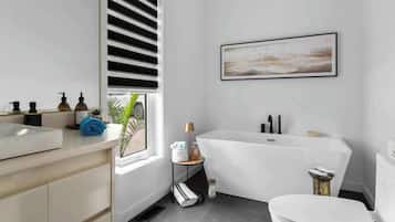 Une salle de bain moderne avec une baignoire, des toilettes et un lavabo. On y trouve une fenêtre avec des stores, un porte-serviettes et une petite table avec une lampe.
