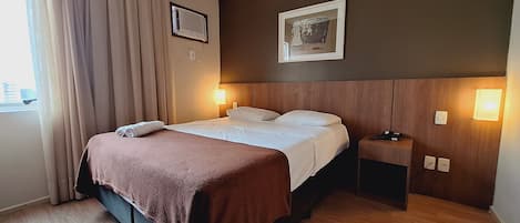 A hotel room with a bed, bedside table, wall-mounted light, and a window with curtains.