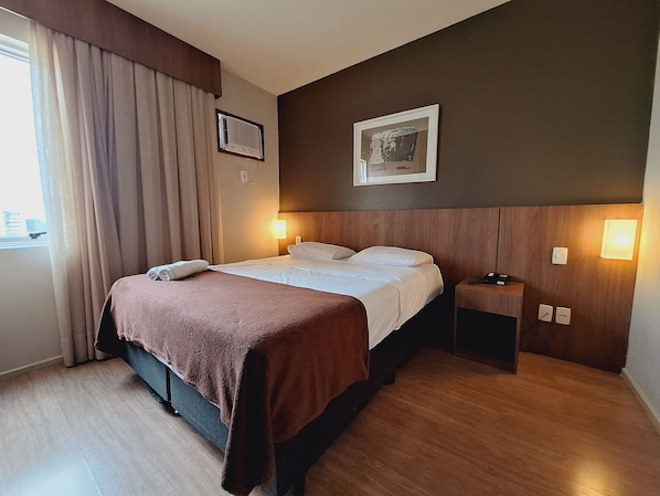 Una habitación de hotel con cama, mesita de noche, lámpara de pared y ventana con cortinas.