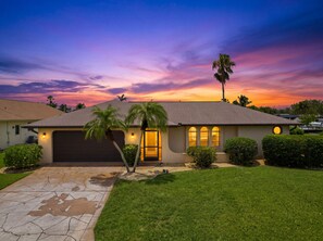 A single-story house with a garage, a well-maintained lawn, and a sunset sky. - Gulf Access Canal Home, Screened Pool | Sleeps 6 (Cape Coral)