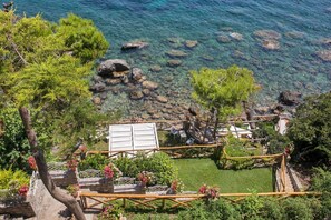 A coastal garden with clear water, rocks, and a wooden deck with seating. - Villa Infinity by My Villa Collection (Massa Lubrense)