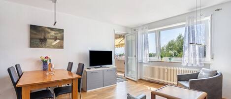 A modern living room with a wooden dining table, grey sofa, and a television on a cabinet.