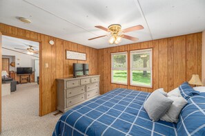 A bedroom with a bed, dresser, television, ceiling fan, and a window with a view of greenery.