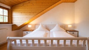 Un dormitorio con techo inclinado, una cama con ropa de cama blanca, dos mesitas de noche con lámparas y una ventana.