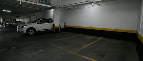 Un estacionamiento subterráneo con pisos de concreto, espacios delimitados y un automóvil blanco estacionado en uno de ellos.