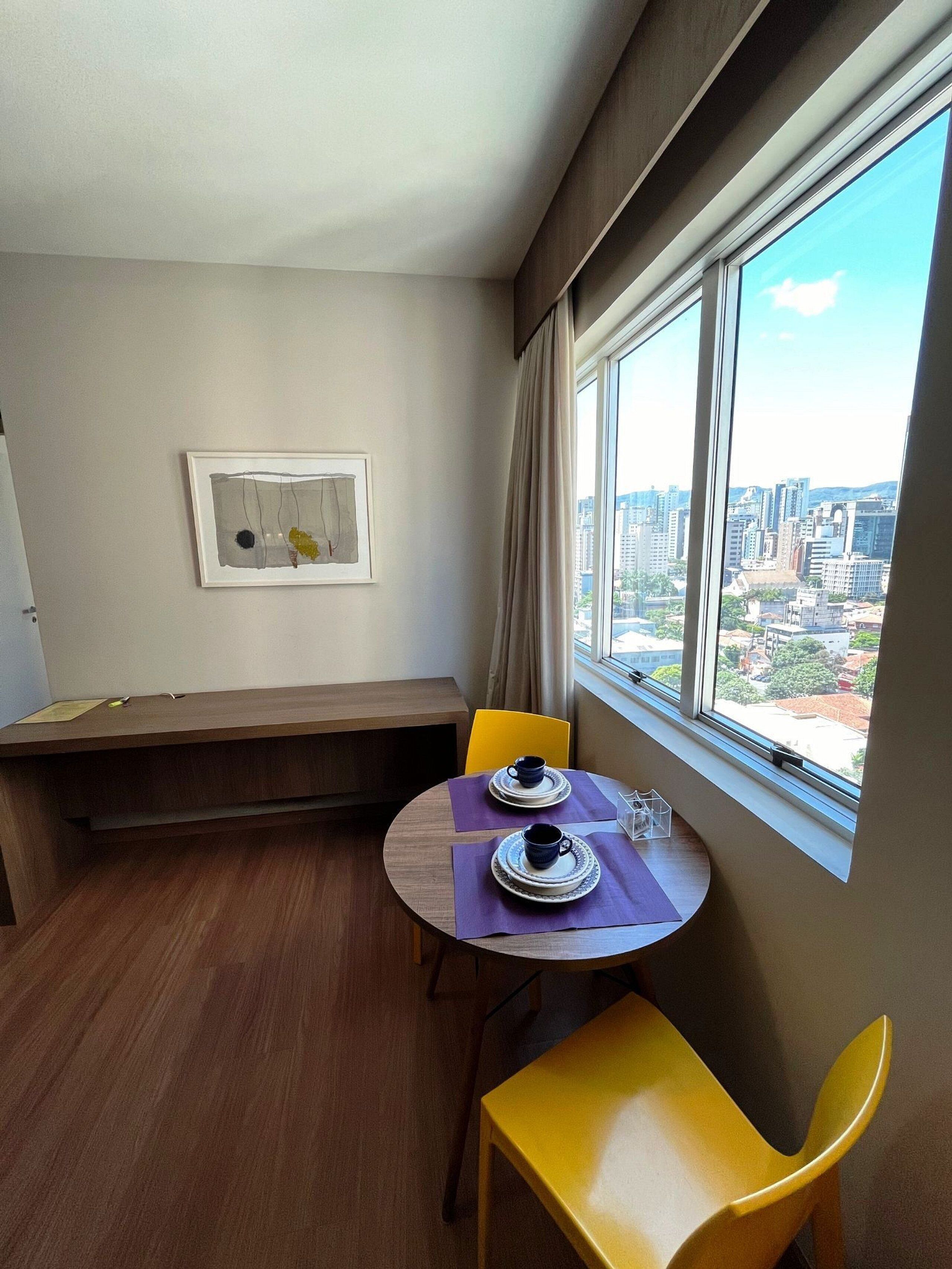 A corner room with a dining table, two chairs, a bench, a window with a city view, and a framed artwork on the wall.