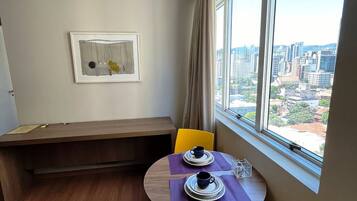 A corner room with a dining table, two chairs, a bench, a window with a city view, and a framed artwork on the wall.