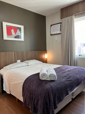 A hotel room with a bed, a framed picture on the wall, a window with curtains, and a wall-mounted air conditioner. - LK Barro Preto 12 (Belo Horizonte)
