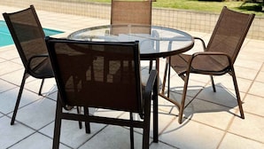 Un área de estar al aire libre con una mesa redonda de vidrio y cuatro sillas, rodeada por un patio con baldosas.