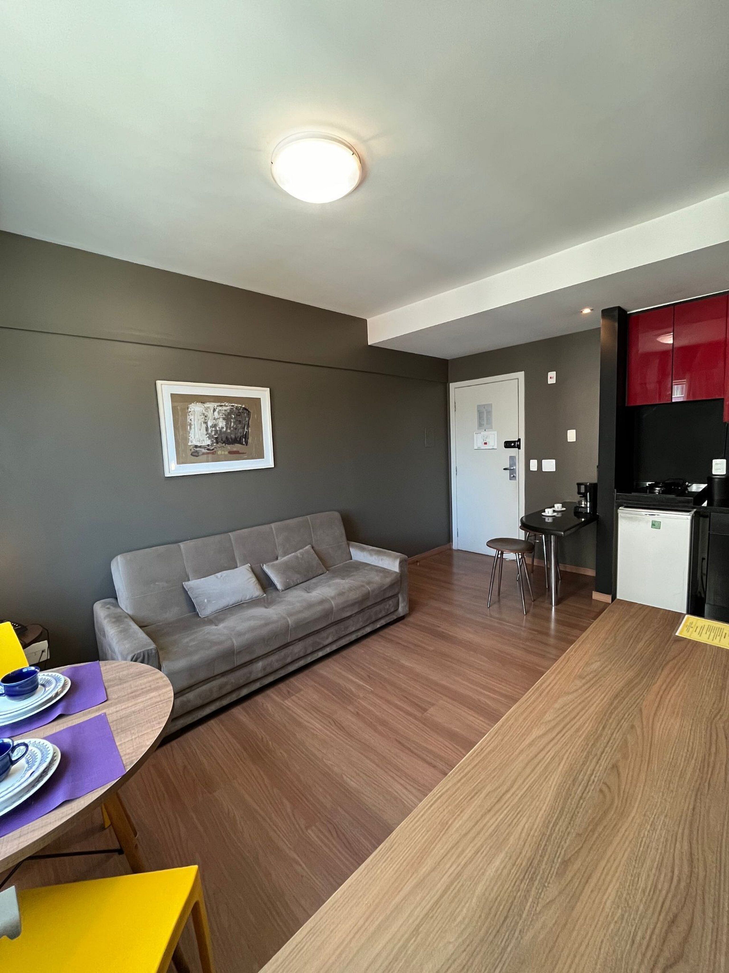 A modern living space with a grey sofa, a wooden dining table, a small kitchen area, and a framed artwork on the wall.