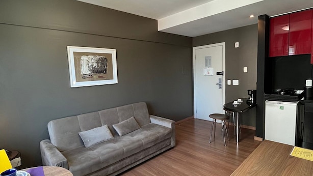 Un espacio habitable moderno con un sofá gris, una mesa de comedor de madera, una pequeña cocina y una obra de arte enmarcada en la pared.