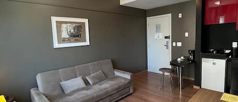 A modern living space with a grey sofa, a wooden dining table, a small kitchen area, and a framed artwork on the wall.