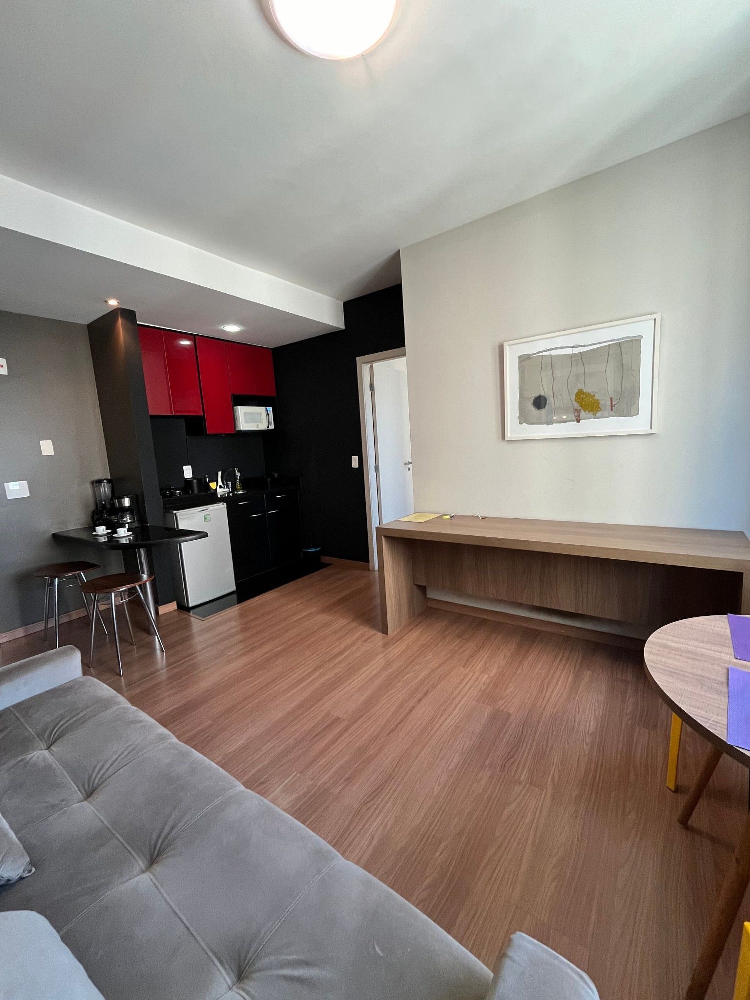 A modern living room with a grey sofa, a wooden coffee table, and a kitchen area with red cabinets and a microwave.