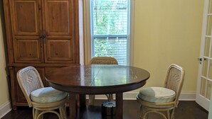 Un coin repas comprenant une table ronde, deux chaises en osier, un buffet en bois et une fenêtre avec des stores.