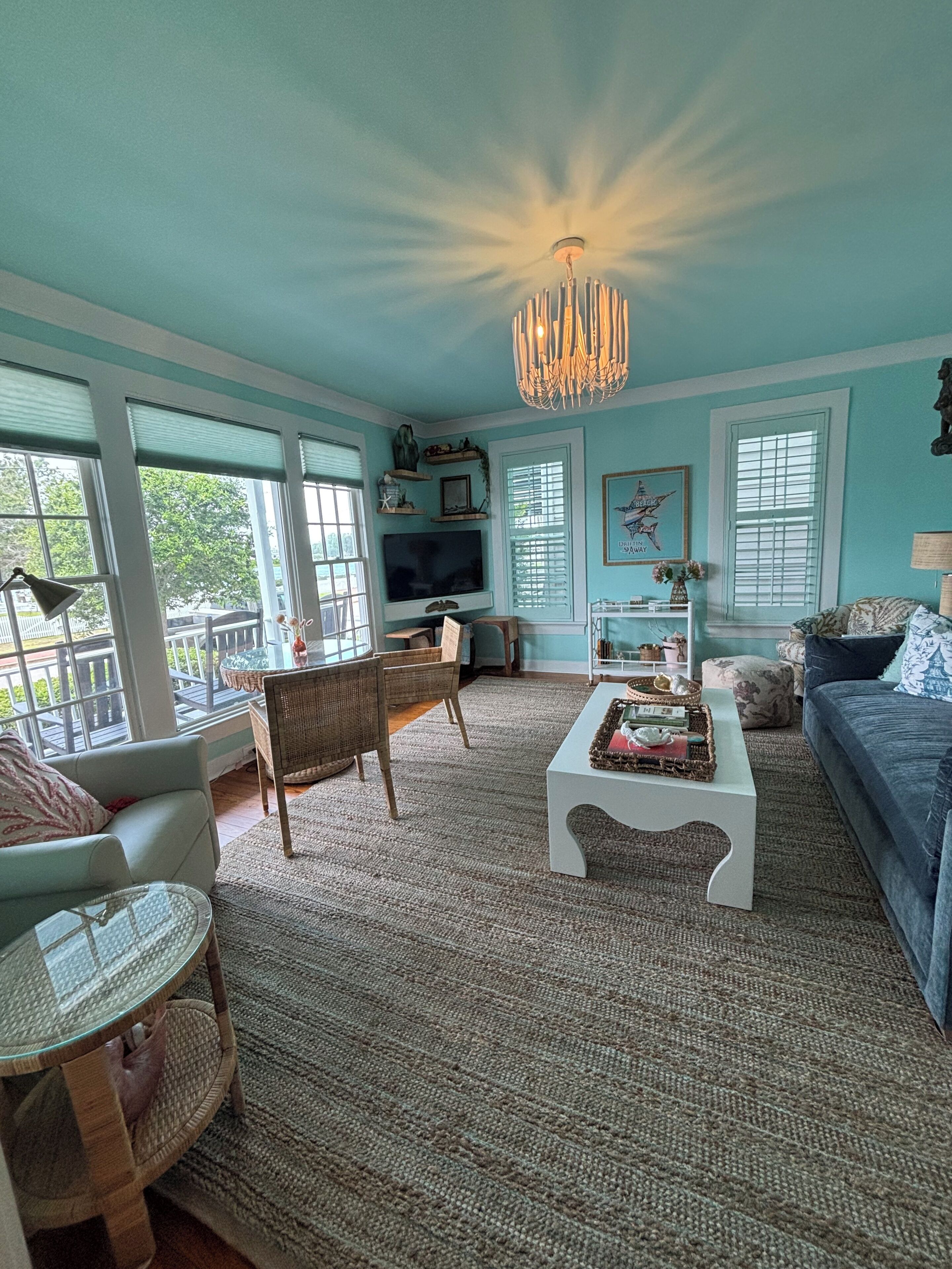 A living room with a television, wicker furniture, a chandelier, and a view through large windows.