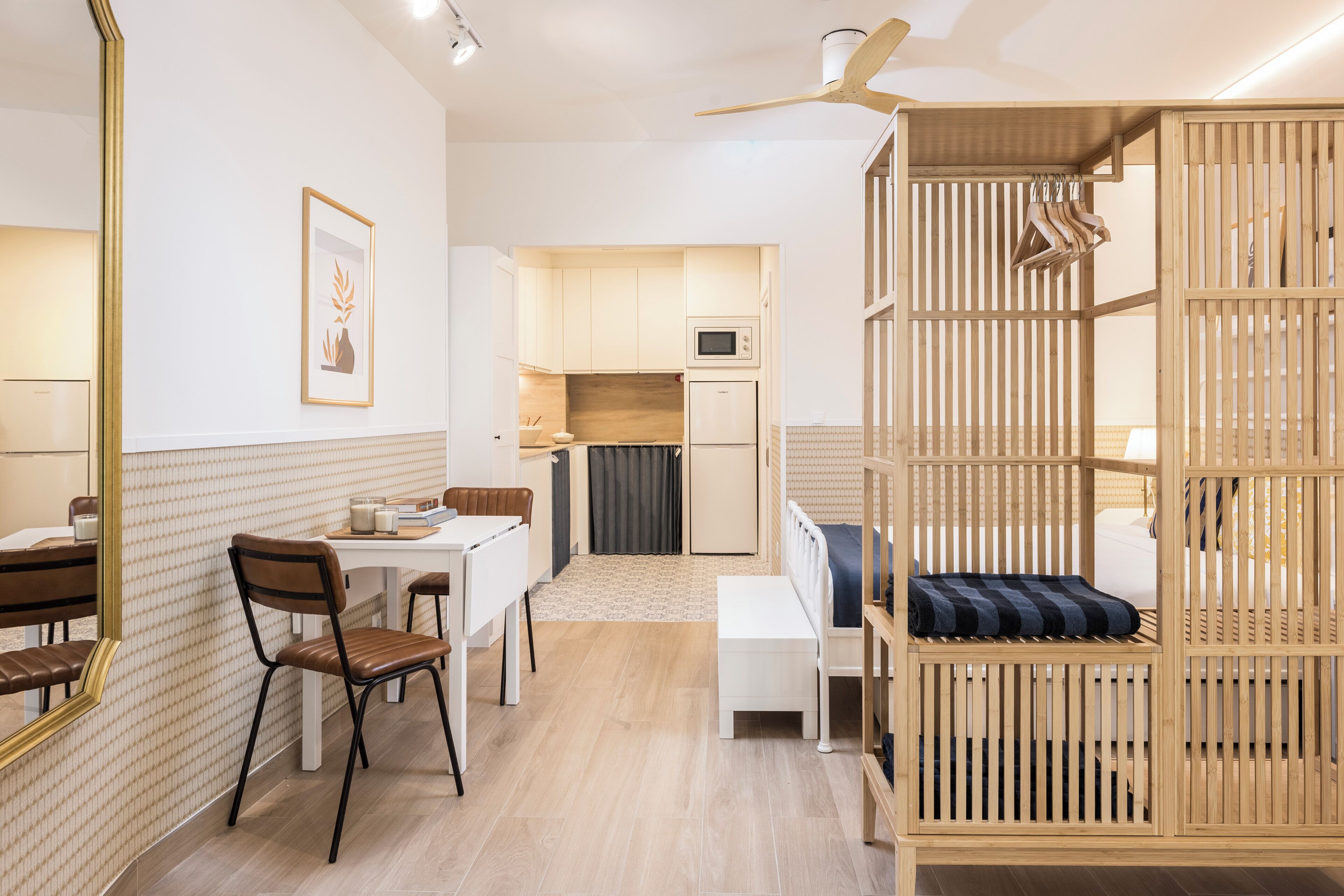 Ein kompaktes Hotelzimmer mit Küchenzeile, Essbereich, einem Bett mit Holz Kopfteil und Deckenventilator.