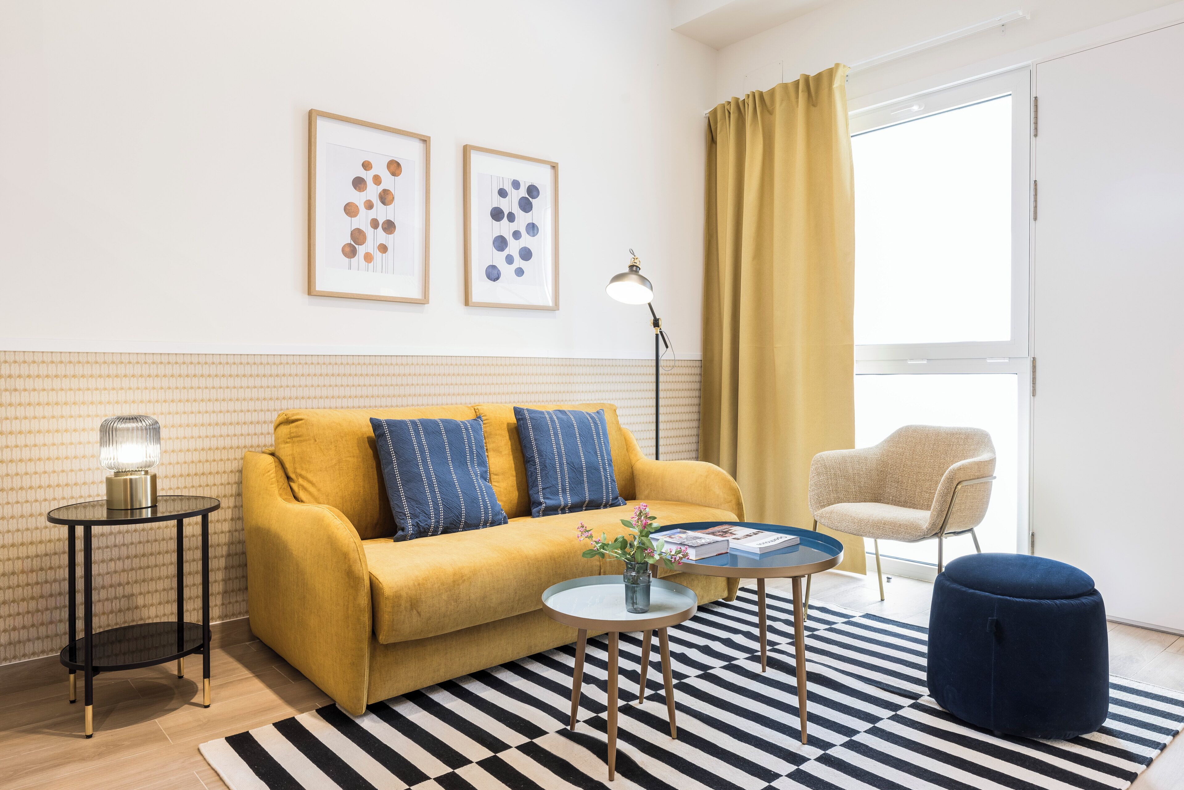 A yellow sofa, a round coffee table, a blue ottoman, a small side table with a lamp, and a chair in a room with a black and white striped rug.