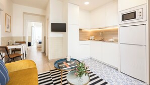 Una cocina moderna con armarios blancos, un microondas y un refrigerador. Hay un comedor con una mesa y sillas, y una sala de estar con un sofá y una mesita de centro.