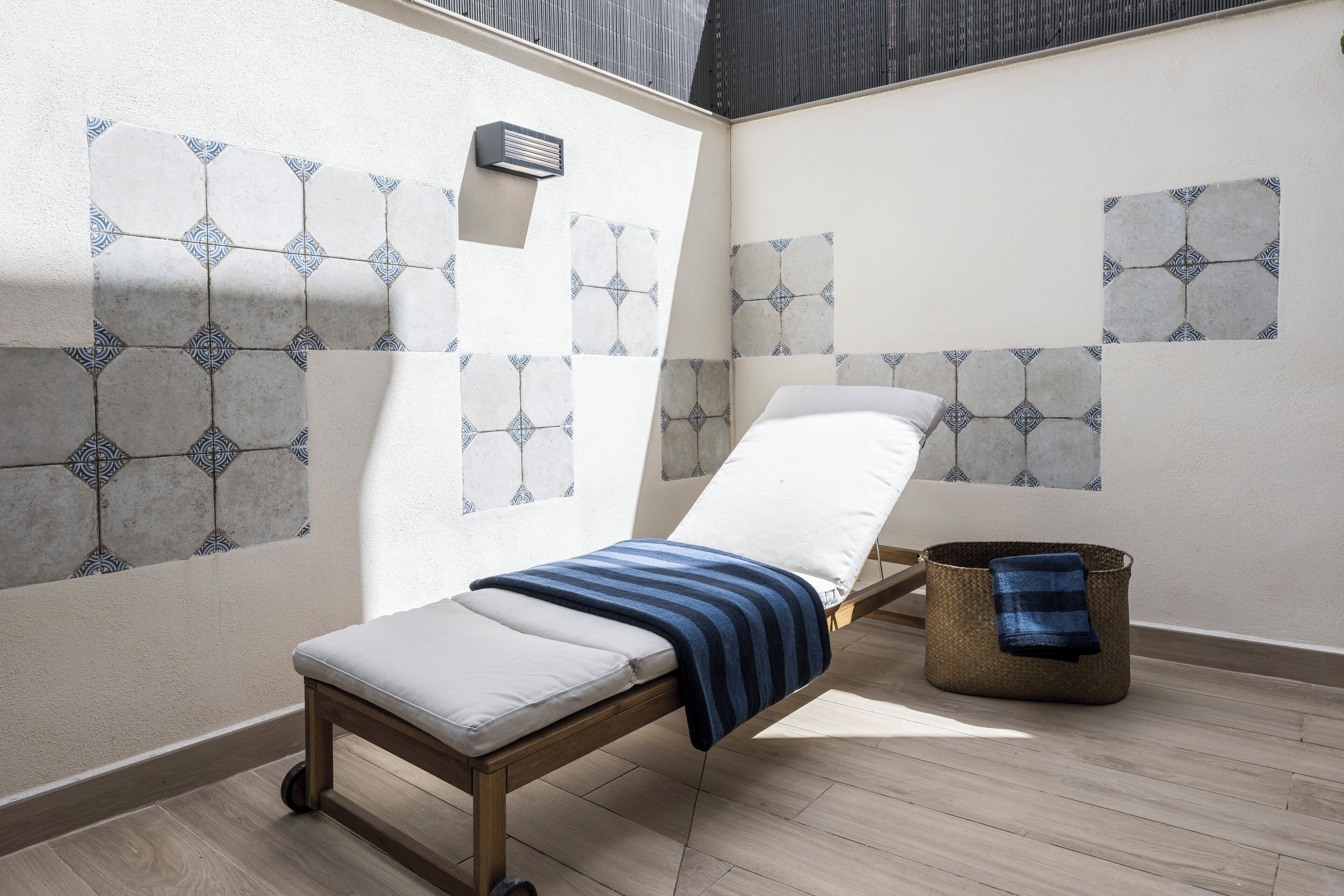 A sunlit room with a sunbed, patterned wall tiles, a small wall-mounted heater, and a woven basket.