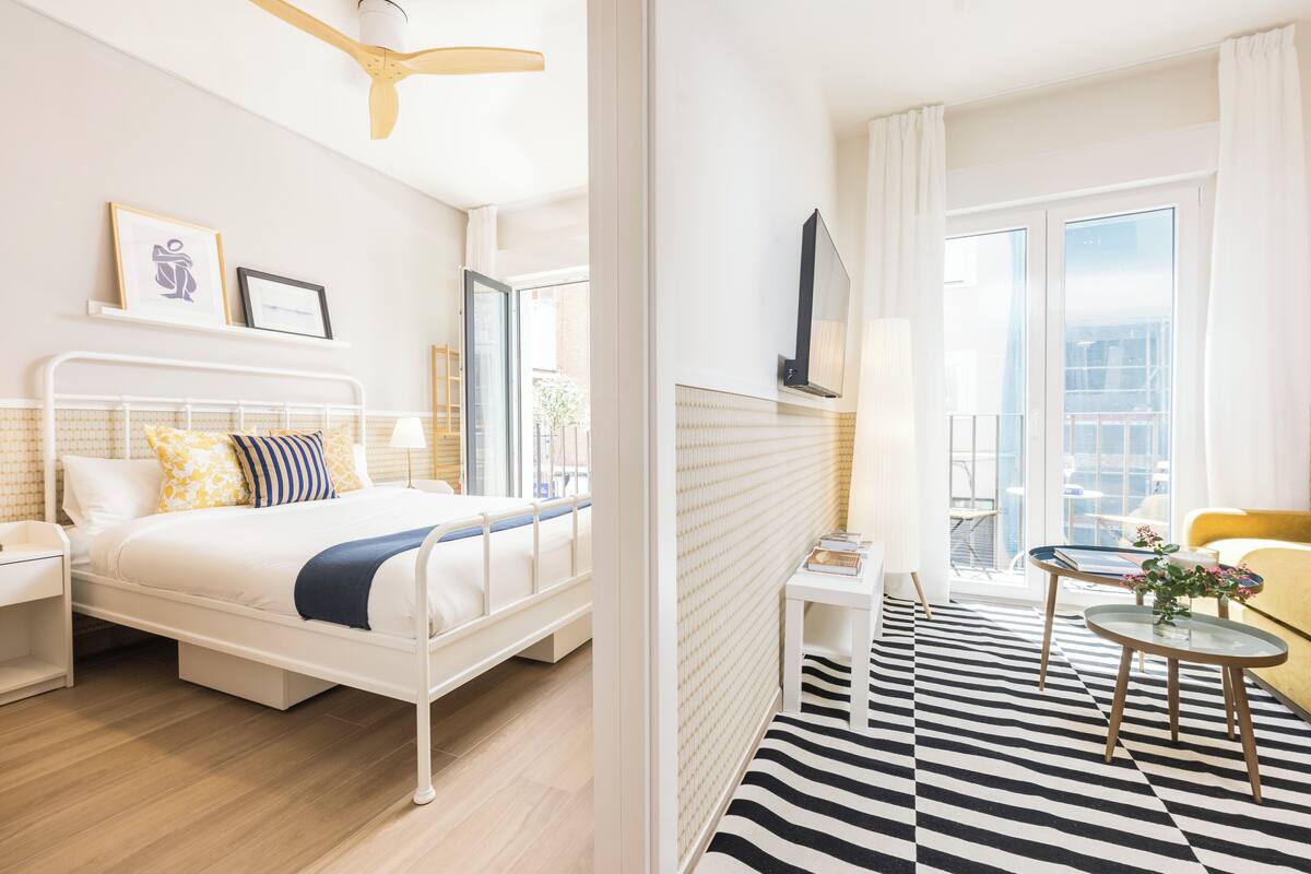 A modern hotel room with a bed, bedside table, lamp, and a view of the city.