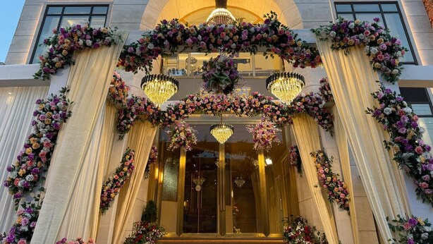 La façade d’un immeuble est ornée d’une enseigne indiquant « SRM » au-dessus de l’entrée, et est flanquée de grandes fenêtres. Des compositions florales et des lustres viennent compléter la décoration.