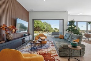 Ein modernes Wohnzimmer mit Sofa, Sesseln, einem Flachbildfernseher und einem großen Fenster mit Blick auf Grünflächen.