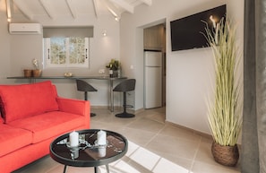 Ein modernes Wohnzimmer mit einem roten Sofa, einem kleinen runden Tisch mit Kerzen, einer Küchenzeile inklusive Kühlschrank und einem an der Wand befestigten Fernseher.